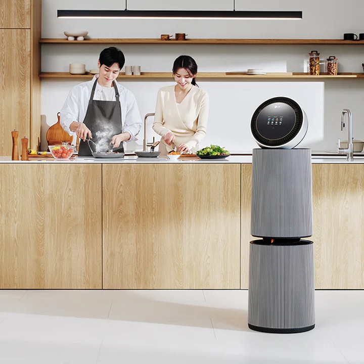 주방 공간에 LG 퓨리케어 AI 오브제컬렉션 360˚ 공기청정기가 있습니다. 나무 재질의 식탁과 의자가 놓여 있고 나무 장식의 주방 가구와 싱크대가 보입니다. 두 사람이 주방에서 요리를 준비하고 있는 모습이 함께 담겨 있습니다.